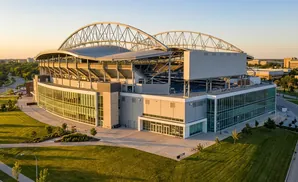 Modern stadium with arched roof at sunset, surrounded by green grass and trees.