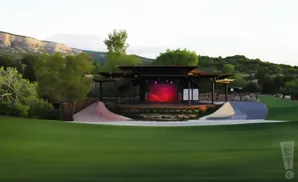 Outdoor amphitheater stage with a red backdrop, set against a landscape of green hills and a rocky cliff under a cloudy sky.