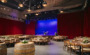 A rustic event space with red stage curtains, banquet tables, and wine barrels.