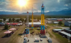 An aerial view of a fairground with tall amusement rides, tents, and mountains in the background.