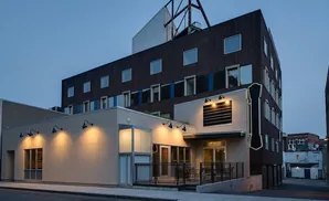 Modern building with dark facade and lighted windows, and a lighter single-story section with outdoor seating.