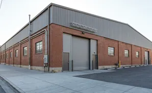 A brick industrial building with a corrugated metal roof and large roll-up doors under a bright sky.