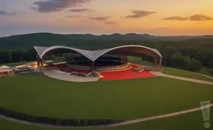 Aerial view of an amphitheater with a curved roof at sunset, surrounded by green hills and trees.