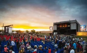 Outdoor concert at sunset with a large crowd seated and standing near a stage.