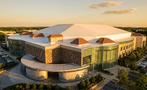 Aerial view of a large, modern building with a white domed roof, curved glass facade, and stone accents during sunset.