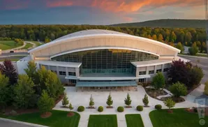 An aerial view of a modern arena with a curved roof, large glass facade, surrounded by trees and green spaces at sunset.