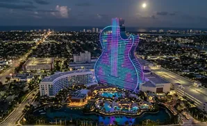Guitar-shaped hotel lit with neon lights at night, overlooking a city and the ocean.