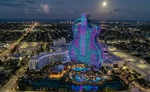 Aerial view of a guitar-shaped hotel lit with neon lights at night, overlooking a city and the ocean.
