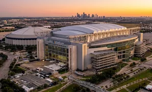 Aerial view of NRG Stadium and Astrodome at sunset, with downtown Houston skyline in the distance.