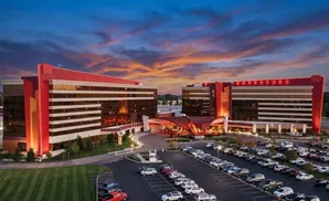Modern casino resort with twin red and glass towers at sunset, illuminated with a parking lot full of cars.