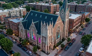 Aerial view of a stone church with stained glass windows illuminated in pink and blue, surrounded by brick buildings and trees.