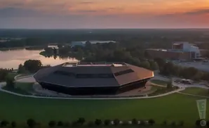 Aerial view of a futuristic, octagonal building on a lake at sunset.
