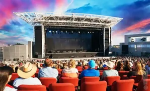 Outdoor concert stage with a large audience seated in red chairs. The sky is a vibrant mix of pink and blue.