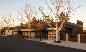 Modern building with wood and brick facade, surrounded by barren trees and landscaping, at sunset.