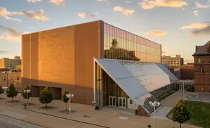 Modern brick and glass building with a dramatic slanted glass entryway, reflecting a cloudy sunset.