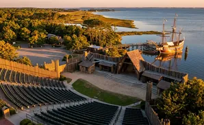An aerial view of a historical reenactment site with a colonial ship docked by a wooden pier, an outdoor amphitheater, and period buildings.
