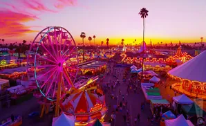 A brightly lit Ferris wheel and carnival rides glow under a vibrant pink and orange sunset sky.