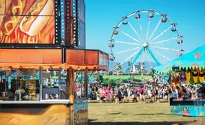 Carnival scene with a Ferris wheel, food stalls, and a crowd of people enjoying the sunny day.