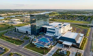 Aerial view of a modern resort with a tall glass building, outdoor pool, and surrounding cityscape.