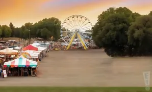 A Ferris wheel and carnival booths are set up at dusk under a colorful sky.