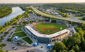 Aerial view of a baseball stadium surrounded by trees and a river, with a highway in the background.