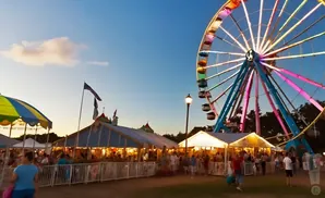 A Ferris wheel illuminates a carnival at dusk, with tents and crowds of people below.