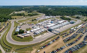 Aerial view of a race track with many cars parked in the paddock area, surrounded by green hills and trees.