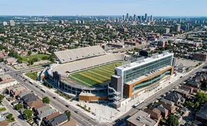 Aerial view of a modern stadium with a glass-fronted building, surrounded by a neighborhood and cityscape.