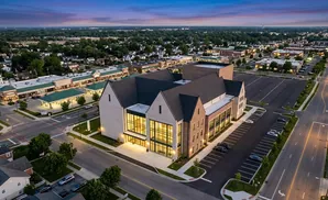 Aerial view of a modern church building with large windows and brick facade, surrounded by a suburban landscape and parking lot at twilight.