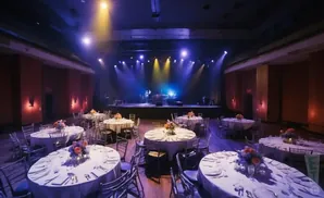 A dimly lit venue with round tables set for an event, facing a stage with a band playing.