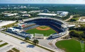 Aerial view of a baseball stadium with surrounding buildings, roads, and trees under a partly cloudy sky.