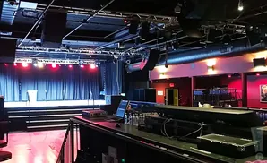 A dimly lit concert venue with a stage, sound equipment, and a bar.