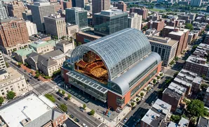 Aerial view of a large glass-domed concert hall with an organ and seating visible inside, surrounded by city buildings.
