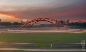 A sunset over a cityscape with a large orange arch sculpture in the foreground.