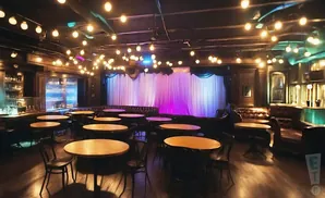 A dimly lit room with round tables and chairs, a bar, and a stage with colorful curtains.