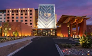 Modern hotel at dusk with a glass-fronted tower, an arched wooden entrance, and warm exterior lighting.