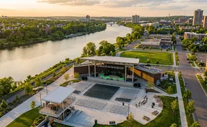 Aerial view of an outdoor amphitheater by a river, with a cityscape in the background at sunset.