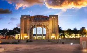 A monumental stone building with arched windows at dusk, illuminated by warm lights under a dramatic sky.