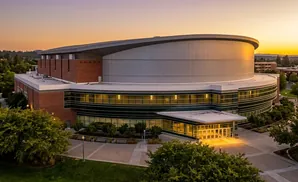 Modern brick and glass arena with a curved metal roof at sunset.
