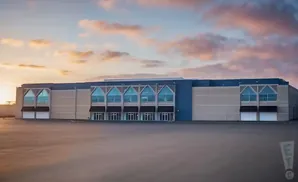 A large building with a beige and blue facade, arched windows, and awnings stands in an empty parking lot under a cloudy sunset sky.