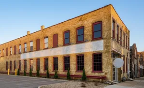 Exterior of a tan brick building with red-trimmed windows, a paved parking lot, and a clear blue sky.