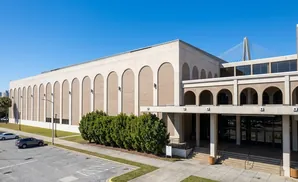 A large, modern building with arched windows and a prominent bridge in the background under a clear blue sky.