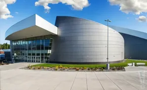 Modern building with curved, silver facade and large glass entrance, under a blue, cloudy sky.