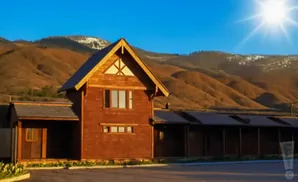 Wooden lodge with snow-capped mountains and a bright sun in a clear blue sky.