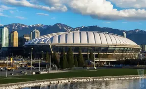 A domed stadium with a white roof and glass walls, set against a backdrop of mountains and city buildings.