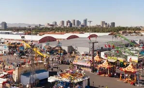 An aerial view of a bustling outdoor carnival with rides, food stands, and a city skyline in the background.