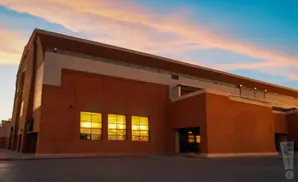 A modern brick building with large windows illuminated from within, set against a sky with soft pink and orange clouds.