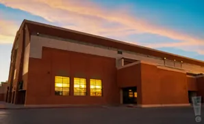 Large brick building with arched roof, lit windows, and a sunset sky.