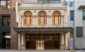 A historic theater with arched windows and an ornate canopy, flanked by modern buildings and a yellow fire hydrant.