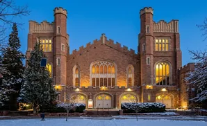 A gothic-style stone building with two towers and arched windows at dusk, surrounded by snow-covered trees.